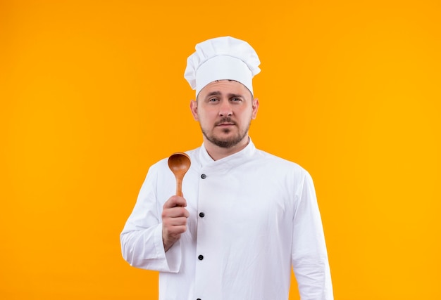 Jeune beau cuisinier confiant en uniforme de chef tenant une cuillère en bois isolée sur un mur orange