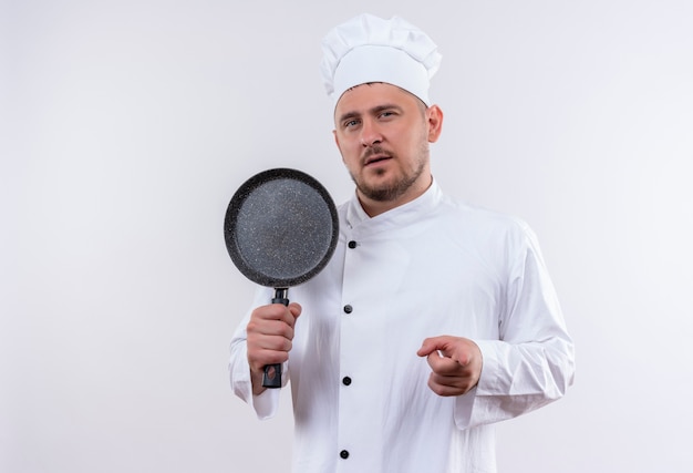 Jeune Beau Cuisinier Confiant En Uniforme De Chef Tenant Une Poêle à Frire Et Pointant Sur Un Mur Blanc Isolé Avec Espace De Copie