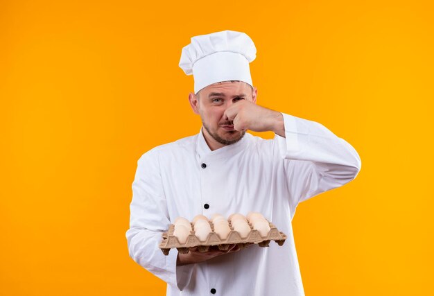 Jeune beau cuisinier irrité en uniforme de chef tenant un carton d'œufs et son nez isolé sur un mur orange