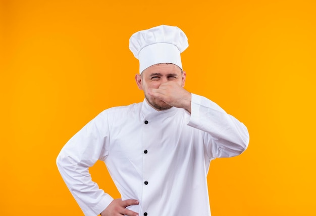 Jeune beau cuisinier irrité en uniforme de chef tenant son nez avec une autre main sur la taille sur un mur orange isolé