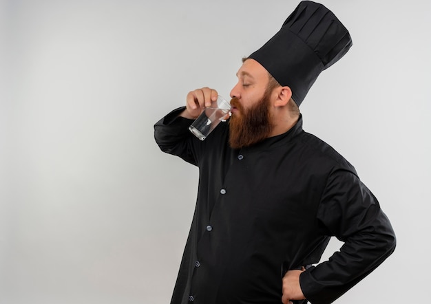 Jeune beau cuisinier en uniforme de chef de l'eau potable du verre avec la main sur la taille et les yeux fermés isolés sur l'espace blanc