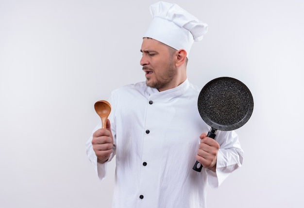 Jeune beau cuisinier en uniforme de chef faire semblant de chanter et de tenir une cuillère et une poêle à frire sur un espace blanc isolé