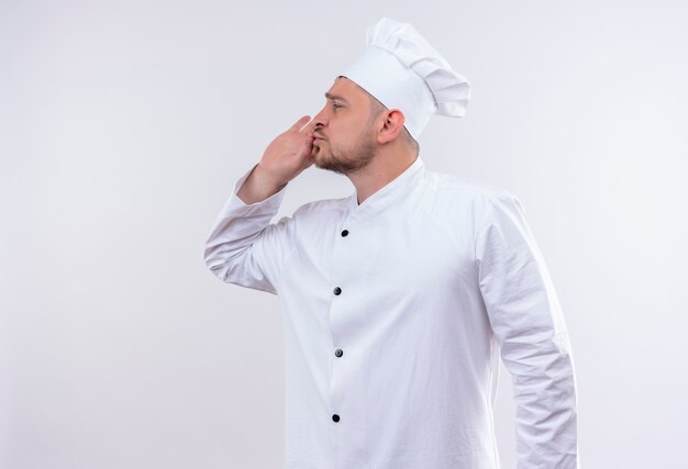 Jeune beau cuisinier en uniforme de chef faisant un geste savoureux à côté sur un espace blanc isolé