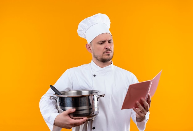 Jeune beau cuisinier en uniforme de chef tenant la chaudière et le bloc-notes et regardant le bloc-notes sur l'espace orange isolé