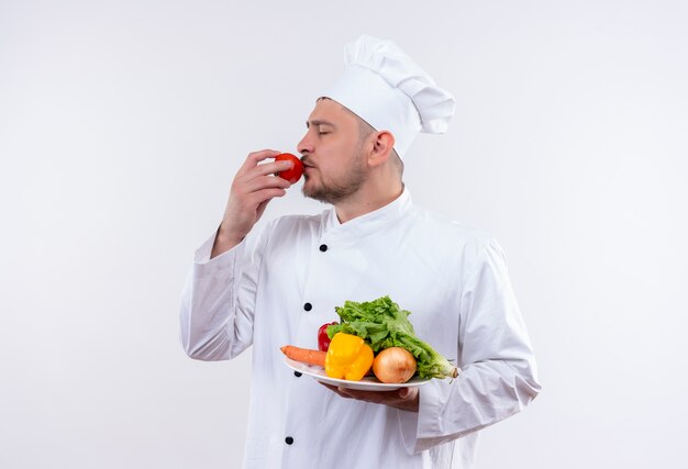 Jeune beau cuisinier en uniforme de chef tenant la plaque avec des légumes et mettre la tomate sur la bouche avec les yeux fermés isolé sur un espace blanc