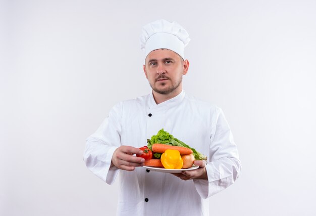 Jeune beau cuisinier en uniforme de chef tenant la plaque avec des légumes à la recherche sur l'espace blanc isolé