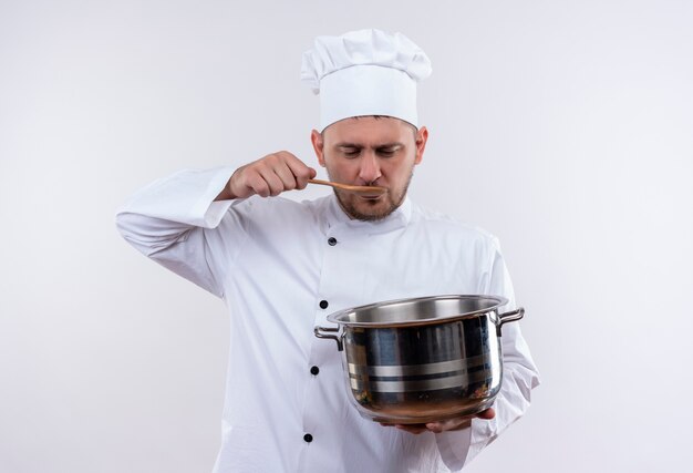 Jeune beau cuisinier en uniforme de chef tenant le pot et mettre une cuillère dans la bouche isolé sur un espace blanc