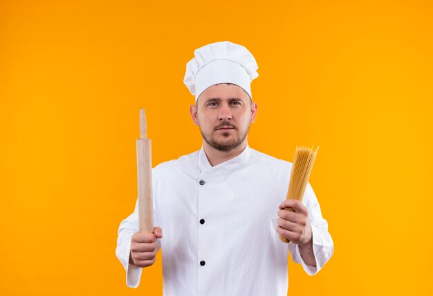 Jeune beau cuisinier en uniforme de chef tenant le rouleau à pâtisserie et les pâtes spaghetti à isolé sur l'espace orange