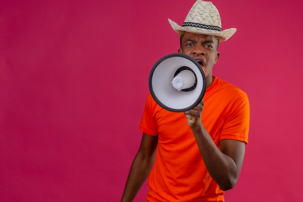 Photo gratuite jeune beau garçon en chapeau d'été portant un t-shirt orange criant au mégaphone avec une expression de colère debout sur un mur rose