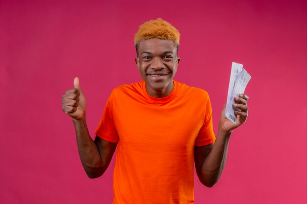 Jeune beau garçon portant un t-shirt orange souriant sorti et heureux tenant des billets d'avion