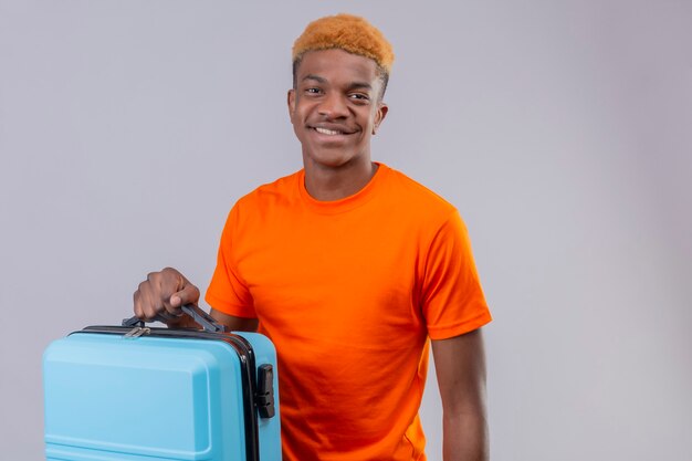 Jeune beau garçon portant un t-shirt orange tenant une valise de voyage souriant positif et heureux debout sur un mur blanc