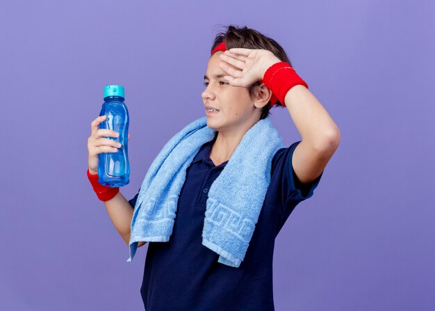 Jeune beau garçon sportif portant un bandeau et des bracelets avec un appareil dentaire et une serviette autour du cou isolé sur un mur violet