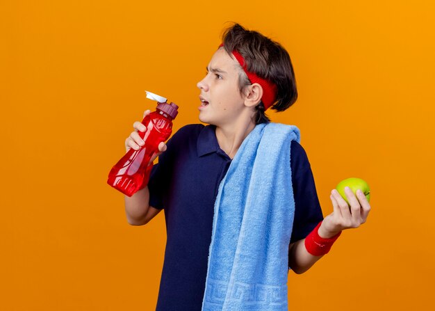 Jeune beau garçon sportif portant un bandeau et des bracelets avec un appareil dentaire et une serviette sur l'épaule tournant la tête à l'autre tenant une bouteille d'eau et de pomme isolé sur un mur orange avec espace de copie