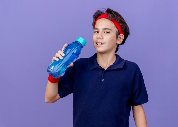 Jeune beau garçon sportif portant un bandeau et des bracelets avec un appareil dentaire tenant une bouteille d'eau regardant la caméra se préparant à boire de l'eau isolé sur fond violet avec espace de copie