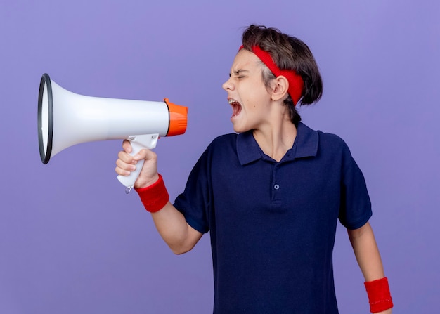 Jeune beau garçon sportif portant un bandeau et des bracelets avec un appareil dentaire tournant la tête à l'autre en criant dans le haut-parleur avec les yeux fermés isolé sur le mur violet