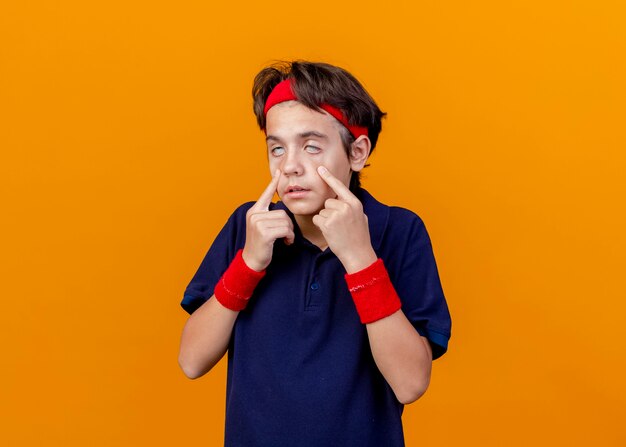 Jeune beau garçon sportif portant un bandeau et des bracelets avec des appareils dentaires tirant vers le bas des paupières isolées sur un mur orange