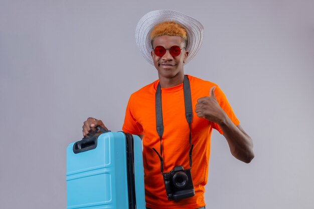 Jeune beau garçon de voyageur en chapeau d'été portant un t-shirt orange tenant une valise de voyage souriant sympathique montrant les pouces debout sur un mur blanc