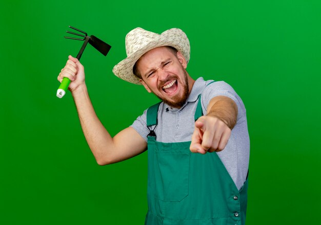 Jeune beau jardinier slave en uniforme et chapeau à la recherche et pointant tenant houe-râteau et crier