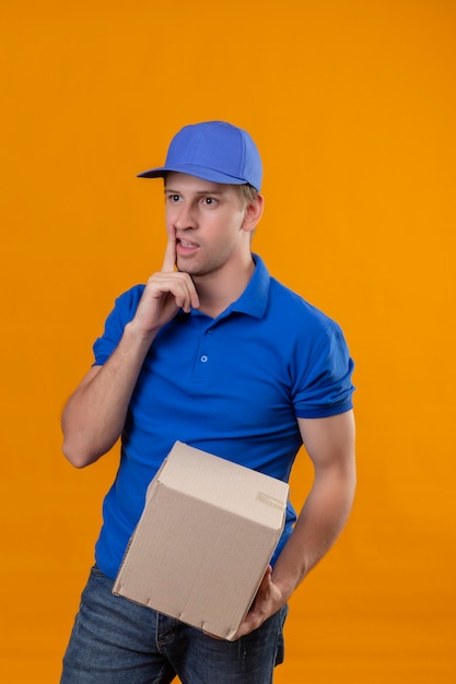 Jeune beau livreur en uniforme bleu et cap holding box package regardant de côté avec une expression pensive sur le visage pensant avoir des doutes debout sur le mur orange