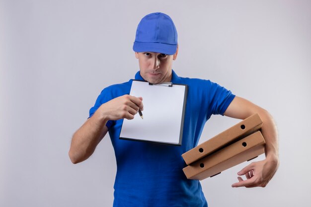 Jeune beau livreur en uniforme bleu et cap tenant des boîtes à pizza et presse-papiers avec des blancs demandant la signature debout sur un mur blanc