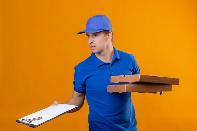 Jeune beau livreur en uniforme bleu et cap tenant des boîtes à pizza et presse-papiers à la confusion et très anxieux debout sur le mur orange