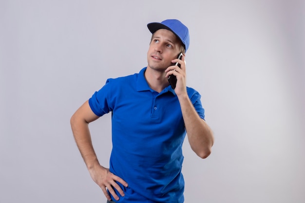 Jeune beau livreur en uniforme bleu et casquette parlant au téléphone mobile avec une expression pensive sur le visage debout sur un mur blanc