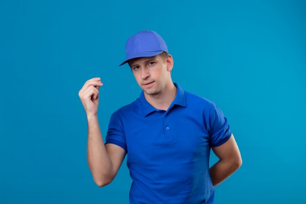 Jeune beau livreur en uniforme bleu et casquette avec visage sérieux faisant le geste de l'argent avec la main debout sur le mur bleu