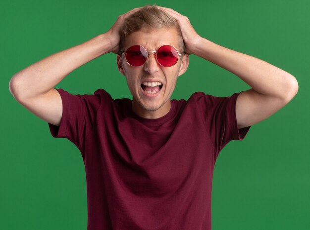Jeune beau mec en colère portant une chemise rouge et des lunettes a attrapé la tête isolée sur le mur vert