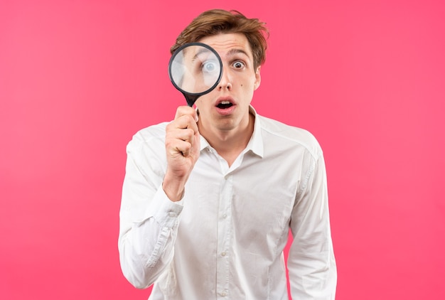 Photo gratuite jeune beau mec confus portant une chemise blanche regardant la caméra avec une loupe