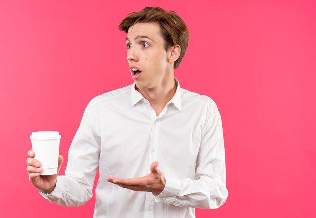 Jeune beau mec confus portant une chemise blanche tenant et pointe avec la main une tasse de café isolée sur un mur rose