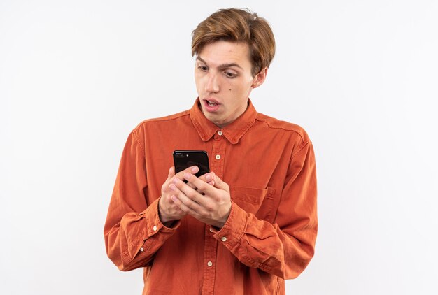 Jeune beau mec confus portant une chemise rouge tenant et regardant le téléphone