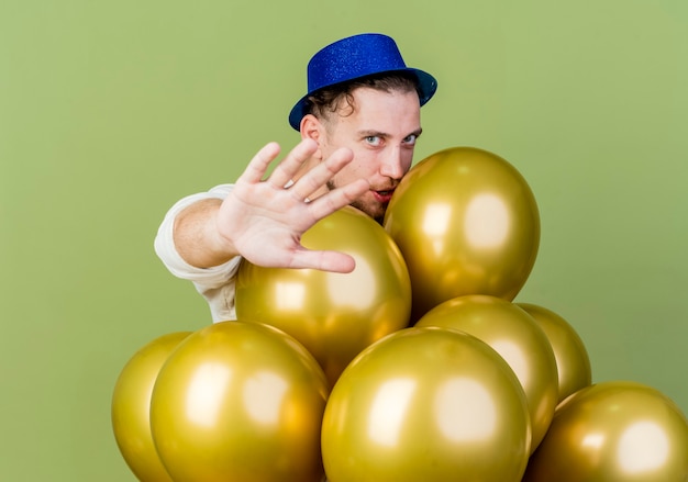 Jeune Beau Mec De Fête Slave Portant Chapeau De Fête Debout Derrière Des Ballons Regardant La Caméra étirant La Main Vers La Caméra Faisant Le Geste D'arrêt Isolé Sur Fond Vert Olive