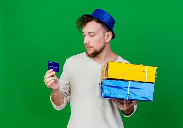 Jeune beau mec de fête slave portant chapeau de fête tenant des coffrets cadeaux et carte de crédit avec les yeux fermés isolé sur fond vert avec espace copie