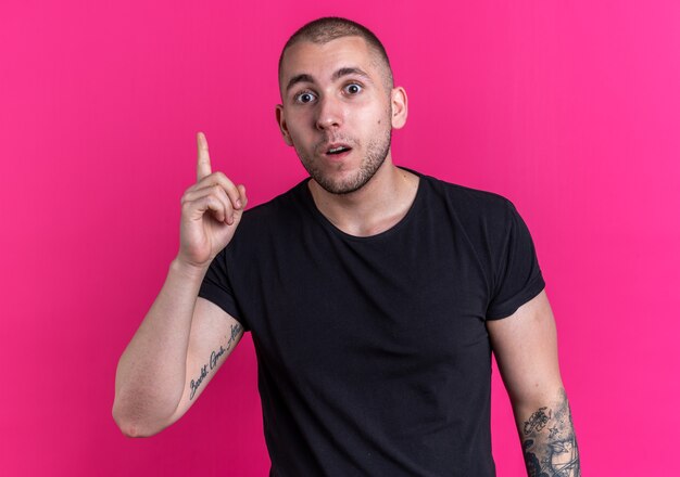 Un jeune beau mec impressionné portant un t-shirt noir pointe vers le haut isolé sur un mur rose