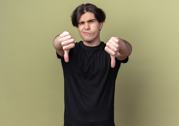 Jeune beau mec mécontent portant un t-shirt noir montrant les pouces vers le bas isolé sur un mur vert olive