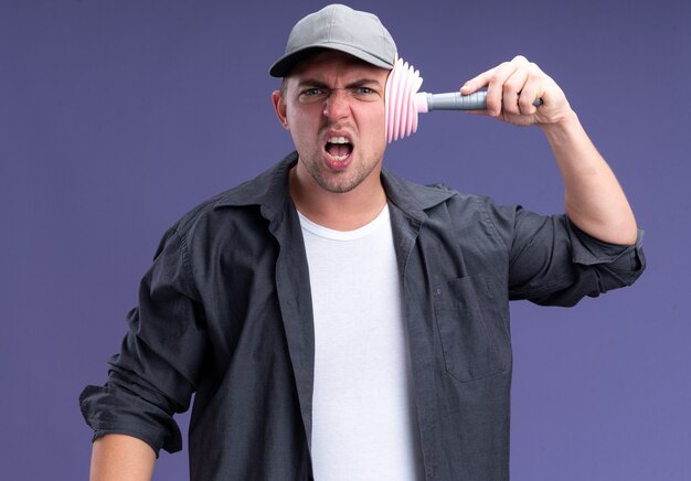 Jeune beau mec de nettoyage en colère portant un t-shirt et une casquette tenant le piston sur la tête isolée sur le mur violet