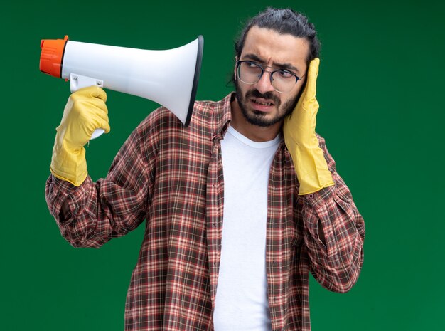 Photo gratuite jeune beau mec de nettoyage concerné portant un t-shirt et des gants tenant et regardant l'oreille couverte de haut-parleur avec la main isolée sur un mur vert