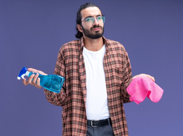 Jeune beau mec de nettoyage confus portant un t-shirt tenant un flacon pulvérisateur avec un chiffon écartant les mains isolées sur un mur bleu