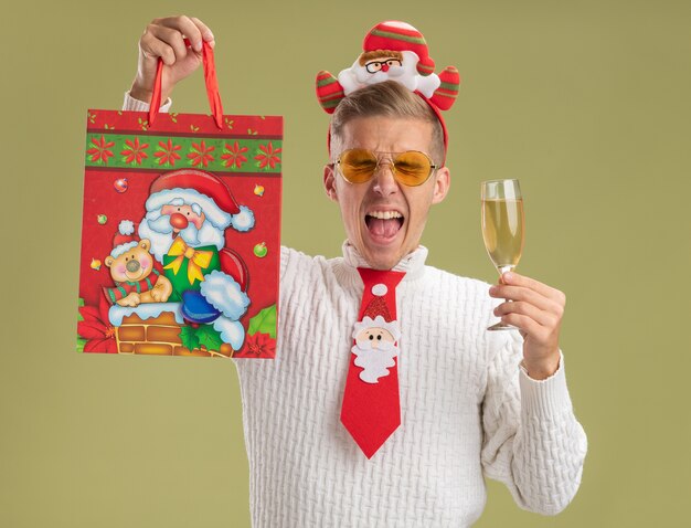 Jeune beau mec portant bandeau et cravate du père noël regardant la caméra tenant un verre de champagne et soulevant le sac de cadeau de Noël et criant les yeux fermés isolé sur fond vert olive