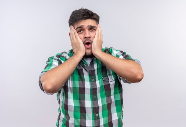 Jeune beau mec portant une chemise à carreaux choqué tenant son visage avec les mains debout sur un mur blanc