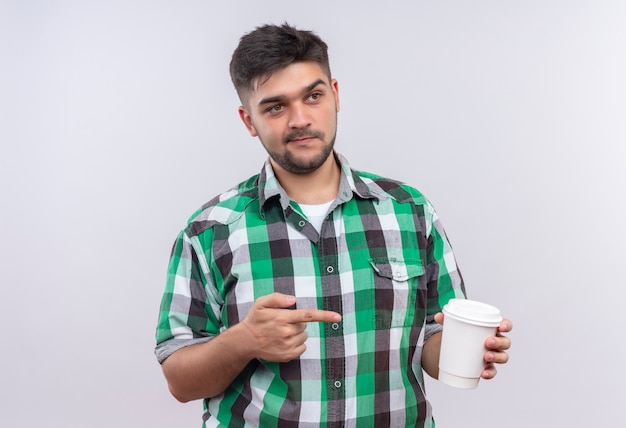 Jeune beau mec portant une chemise à carreaux à l'évaluation en plus de pointer vers une tasse de café plastik debout sur un mur blanc