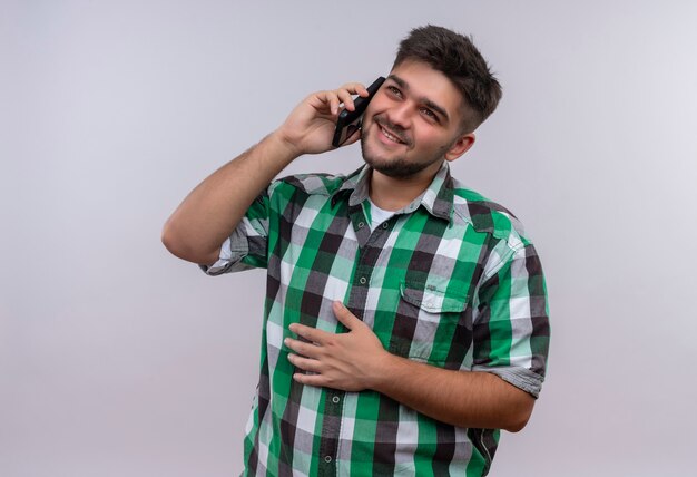 Jeune beau mec portant une chemise à carreaux joyeusement regardant parler au téléphone debout sur un mur blanc