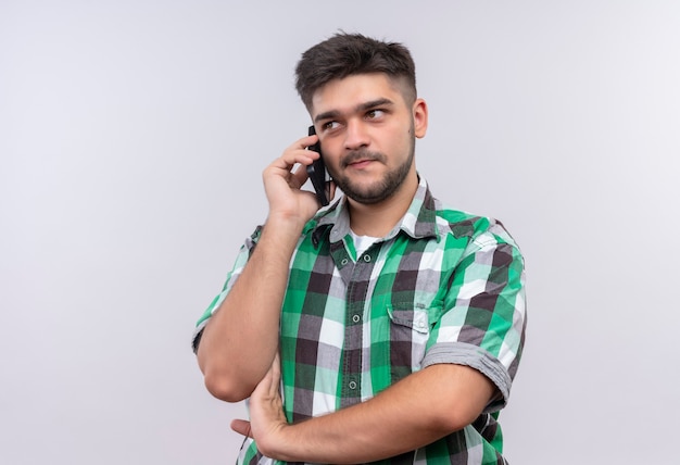 Photo gratuite jeune beau mec portant une chemise à carreaux parlant au téléphone à la recherche en plus de se tenir debout sur un mur blanc