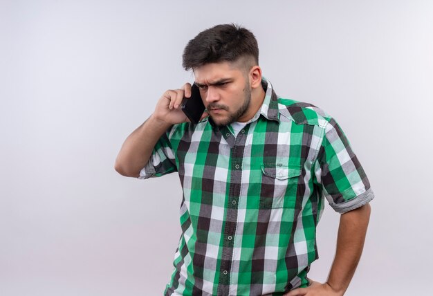 Jeune beau mec portant une chemise à carreaux parler avec colère au téléphone debout sur un mur blanc