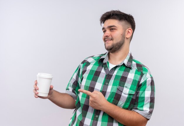 Jeune beau mec portant une chemise à carreaux en plus heureux de pointer vers une tasse de café plastik debout sur un mur blanc