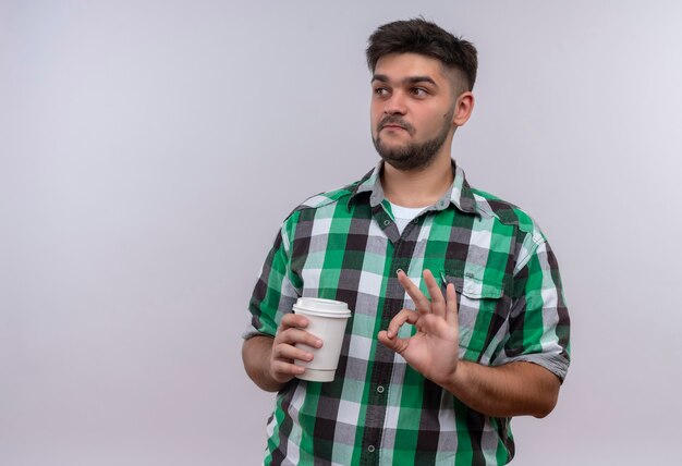 Jeune beau mec portant une chemise à carreaux en plus de louer le café debout sur un mur blanc