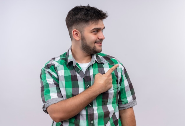 Jeune beau mec portant une chemise à carreaux en plus de pointer vers la gauche avec l'index debout sur un mur blanc