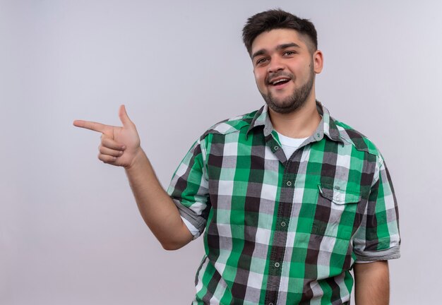 Jeune beau mec portant une chemise à carreaux à pointant joyeusement vers la droite avec l'index debout sur un mur blanc