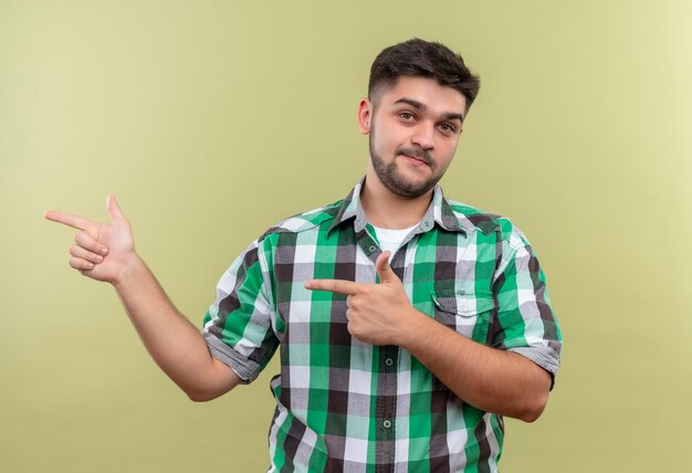 Jeune beau mec portant une chemise à carreaux pointant vers la droite avec l'index debout sur un mur kaki