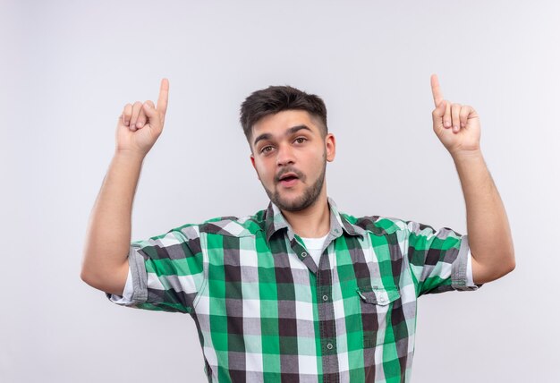 Jeune beau mec portant une chemise à carreaux pointant vers le haut avec l'index debout sur un mur blanc